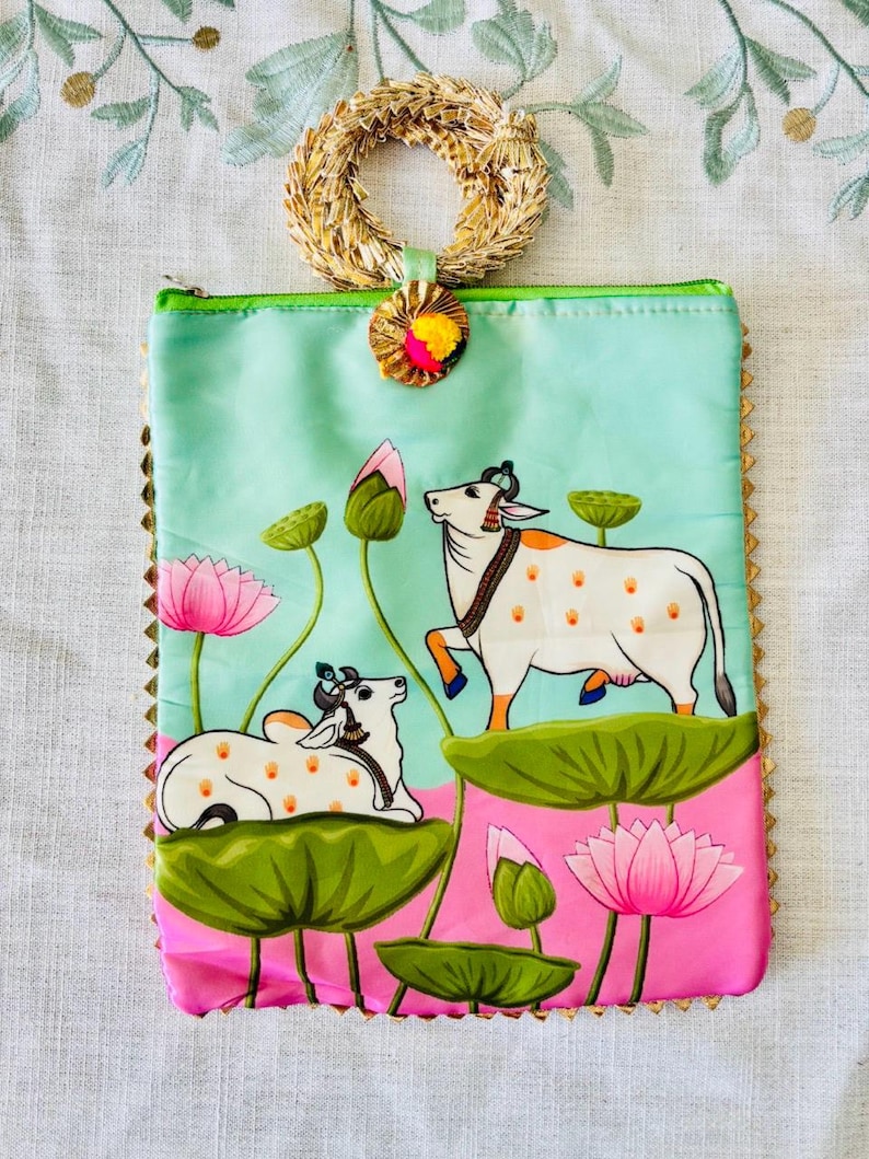Pode incluir: Uma bolsa retangular com al&ccedil;a tecida e ilustra&ccedil;&atilde;o colorida. A bolsa apresenta um fundo azul claro e rosa com uma pintura de duas vacas brancas, flores de l&oacute;tus e folhas de nen&uacute;far verdes. A bolsa &eacute; adornada com dourado e tem um z&iacute;per verde.