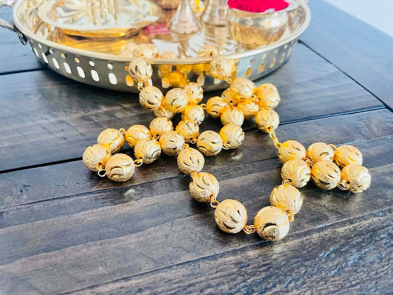 May include: A gold beaded garland with intricate designs on each bead. The garland is made of metal and has a shiny finish.