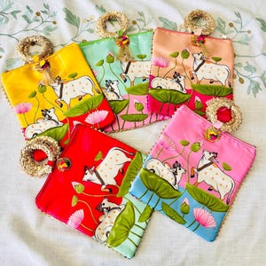 Pode incluir: Uma cole&ccedil;&atilde;o de bolsas de tecido quadradas e coloridas. Cada bolsa apresenta um design pintado &agrave; m&atilde;o de uma vaca branca com manchas marrons, flores de l&oacute;tus e folhas verdes. As bolsas est&atilde;o em tons de amarelo, vermelho, rosa, verde e p&ecirc;ssego.
