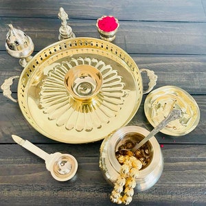 May include: A brass tray with a decorative design, a small bowl, a lidded container, a spoon, and a small bowl with red powder. The tray has a raised edge and two handles. The bowl is in the center of the tray and has a floral design. The lidded container is on the right side of the tray and has a round shape. The spoon is in the lidded container. The small bowl with red powder is on the left side of the tray.