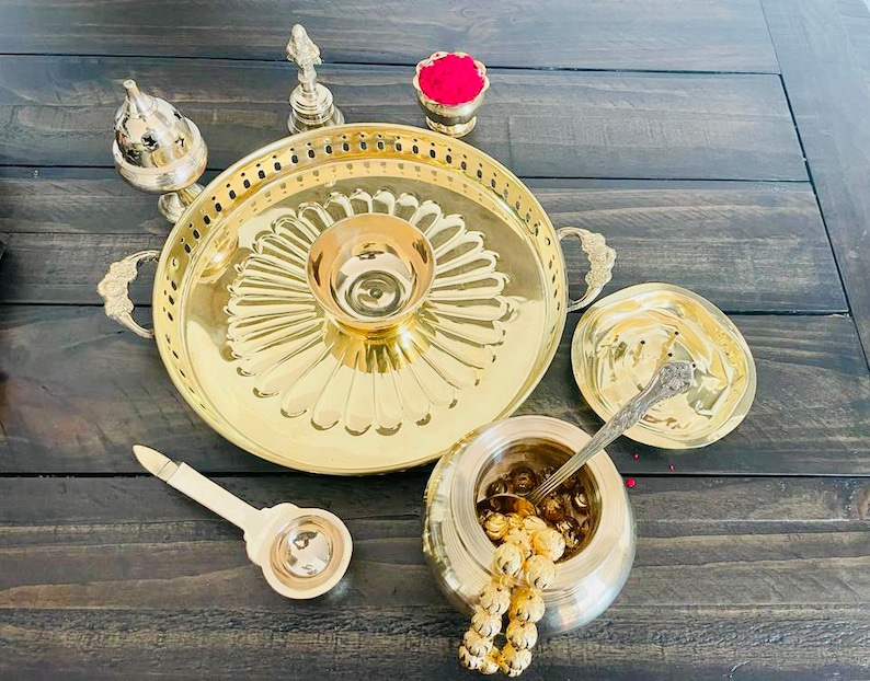 May include: A brass tray with a decorative design, a small bowl, a lidded container, a spoon, and a small bowl with red powder. The tray has a raised edge and two handles. The bowl is in the center of the tray and has a floral design. The lidded container is on the right side of the tray and has a round shape. The spoon is in the lidded container. The small bowl with red powder is on the left side of the tray.