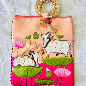 Pode incluir: Uma bolsa de m&atilde;o cor de p&ecirc;ssego com um desenho pintado &agrave; m&atilde;o de duas vacas em nen&uacute;fares, rodeadas por flores de l&oacute;tus rosa e folhas verdes. A bolsa tem uma al&ccedil;a de palha tecida e uma borda dourada.