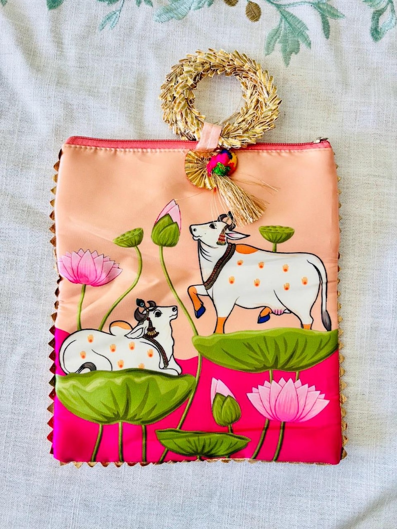 Pode incluir: Uma bolsa de m&atilde;o cor de p&ecirc;ssego com um desenho pintado &agrave; m&atilde;o de duas vacas em nen&uacute;fares, rodeadas por flores de l&oacute;tus rosa e folhas verdes. A bolsa tem uma al&ccedil;a de palha tecida e uma borda dourada.