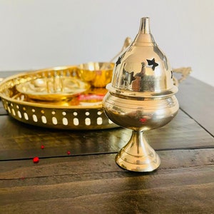 May include: A gold metal incense burner with a star-shaped design. The burner is on a wooden table with a gold metal tray in the background.