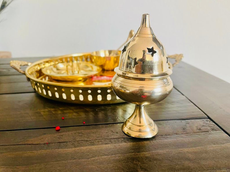 May include: A gold metal incense burner with a star-shaped design. The burner is on a wooden table with a gold metal tray in the background.