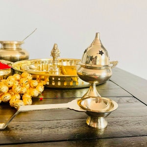 May include: A set of brass puja items, including a plate, a small bowl, a bell, and a incense burner. The items are arranged on a wooden table.