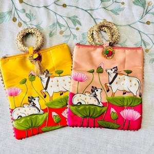 Pode incluir: Duas bolsas de tecido decorativas, uma amarela e outra p&ecirc;ssego. Cada uma apresenta um desenho pintado &agrave; m&atilde;o de vacas brancas, flores de l&oacute;tus cor-de-rosa e folhas de nen&uacute;far verdes. T&ecirc;m al&ccedil;as de anel douradas e borlas decorativas.