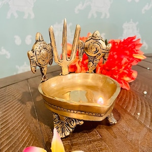 May include: A gold-colored brass oil lamp with a heart-shaped bowl and three turtle legs. The lamp has a trident and a stylized turtle design on the sides. A small flame is burning in the bowl.