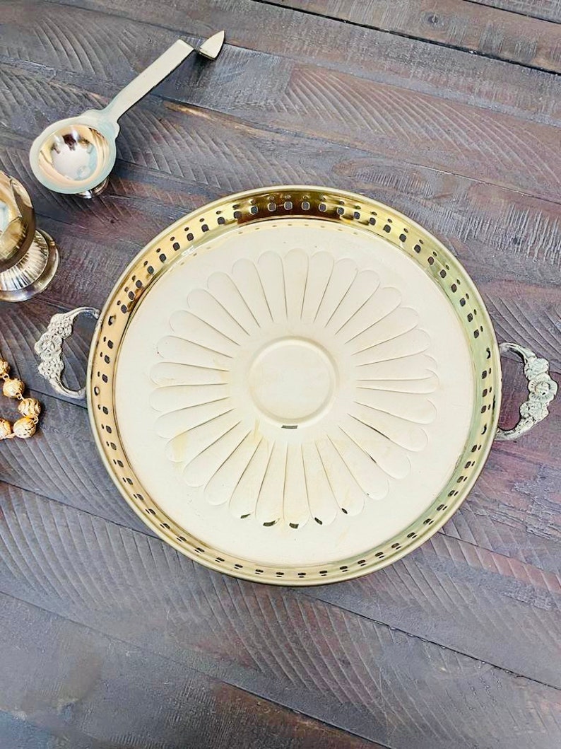 May include: A gold serving tray with a decorative floral design and two handles. The tray is on a wooden surface.