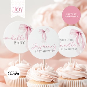 May include: Cupcake toppers for a baby shower in pink and white. The toppers say "hello BABY", "Jasmine's BABY SHOWER", and "SWEET LITTLE bundle IS ON THE WAY". Each has a pink bow design. The cupcakes have pink icing.