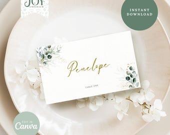 May include: A white place card with a green and gold floral design. The name "Penelope" is written in gold script and the text "Table One" is printed in gray below the name.