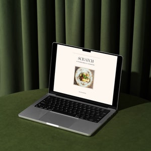 May include: A silver laptop computer open to a webpage with the title "Scratch: A Prometabolic Cookbook". The webpage features a photo of a plate of food. The laptop is on a green surface.