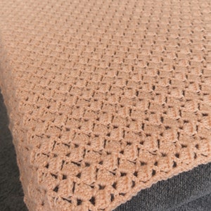 May include: A peach-colored crochet blanket with a textured pattern. The blanket is draped over a gray surface, showcasing the intricate stitch work. The soft, warm tones suggest a cozy, handmade item.
