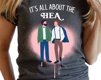 Może przedstawiać: Ciemnoszara koszulka z napisem "IT'S ALL ABOUT THE HEA" nad ilustracją dwóch mężczyzn trzymających się za ręce. Jeden mężczyzna ma na sobie zieloną kurtkę, drugi niebieską pelerynę. Różowy efekt rozprysku znajduje się za ilustracją.