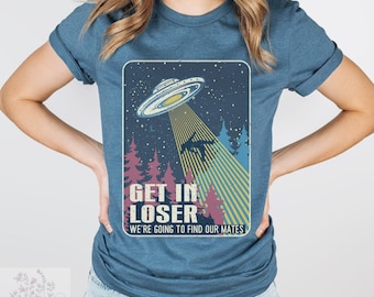 May include: Blue t-shirt featuring a retro-style graphic of a UFO beaming up a figure in a forest. The text "GET IN LOSER WE'RE GOING TO FIND OUR MATES" is printed below the image. The shirt has short sleeves.