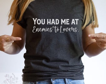 May include: A dark gray t-shirt with the text "You had me at Enemies to Lovers." printed in white.