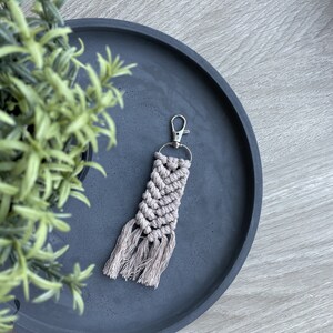 May include: A light brown macrame keychain with a silver key ring and tassels. The keychain is on a dark gray tray.