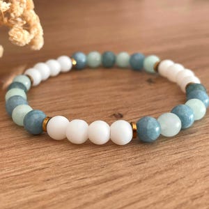 Könnte beinhalten: Ein Armband mit weißen, hellblauen und mintgrünen Perlen. Es hat goldfarbene Abstandshalter zwischen den Perlen. Das Armband liegt auf einer Holzoberfläche.