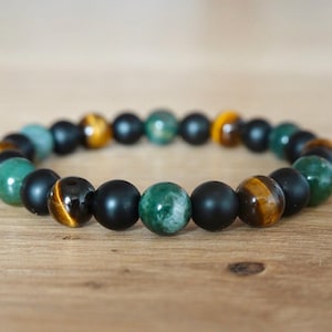 Könnte beinhalten: Ein Armband mit Perlen in verschiedenen Farben, darunter mattes Schwarz, Grün und Tigerauge. Das Armband ist aufgefädelt und zeigt eine Vielzahl von Texturen und Farbtönen. Die Perlen haben einen Durchmesser von etwa 10 mm.