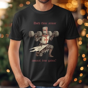 Könnte beinhalten: Schwarzes T-Shirt mit einem Cartoon-Ritter, der Gewichte hebt. Der Ritter trägt eine Rüstung und einen Helm mit rotem Kreuz. Der Text auf dem Shirt lautet "Doth thine armor conceal true gains?"