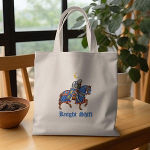 May include: Cream-colored tote bag featuring a medieval knight on horseback with blue and gold accents. The design includes a crescent moon and the words "Knight Shift" in a stylized font. The bag has two cream-colored handles.