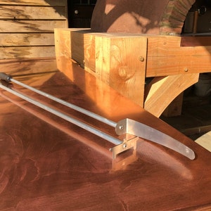 Op de afbeelding: Twee lange, zilveren metalen pizzascheppen met houten handvatten, rustend op een koperen oppervlak. De scheppen zijn ontworpen voor gebruik in een houtoven.