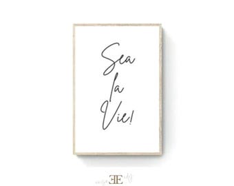 May include: A rectangular wall art print featuring a light wood frame and a white background. The text Sea la Vie! is printed in black in a cursive script font, arranged vertically in three lines. The design is centered within the frame.