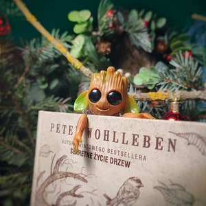 Può includere: Una piccola statuetta di legno marrone con occhi grandi e un sorriso, che tiene un libro intitolato "Pete Wohlleben, Autore del bestseller internazionale, La vita segreta degli alberi".