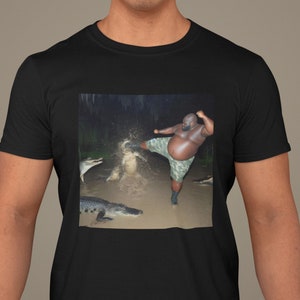 Może przedstawiać: Czarny t-shirt z nadrukiem graficznym przedstawiającym mężczyznę kopiącego krokodyla. Grafika ma kształt kwadratu i jest wyśrodkowana na koszulce. Mężczyzna ma na sobie krótkie spodenki w kamuflażu i kopie krokodyla prawą nogą. Krokodyl jest w wodzie i patrzy na mężczyznę. Tło jest ciemne i woda pryska wokół mężczyzny i krokodyla.