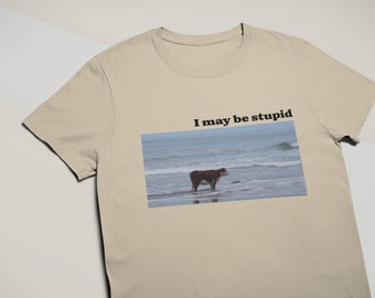 Könnte beinhalten: Ein beiges T-Shirt mit dem Text "I may be stupid" auf der Vorderseite. Der Text ist schwarz und mittig auf dem Shirt platziert. Unter dem Text befindet sich ein Bild einer braunen Kuh, die im Meer steht. Das Meer ist blau und im Hintergrund sind Wellen zu sehen.