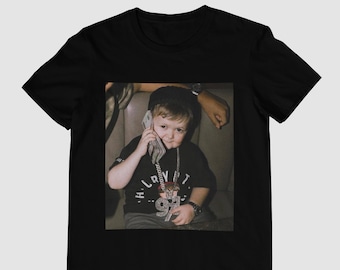 May include: A black t-shirt with a photo of a young boy wearing a large diamond chain and holding a stack of cash to his ear. The boy is sitting in a car and wearing a black shirt with the text "LVIT" on it.
