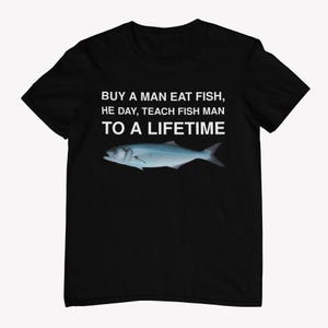 Peut inclure: T-shirt noir avec un texte blanc qui dit "BUY A MAN EAT FISH, HE DAY, TEACH FISH MAN TO A LIFETIME". Un poisson bleu est représenté sous le texte.