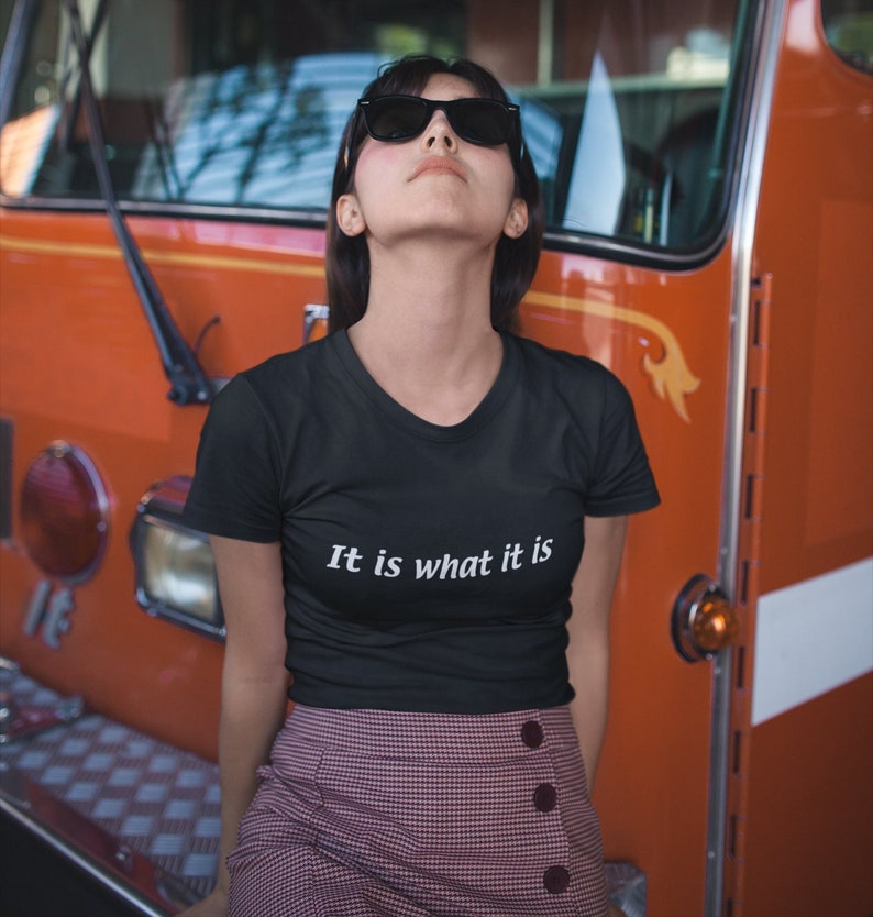K&ouml;nnte beinhalten: Eine Frau tr&auml;gt ein schwarzes T-Shirt mit dem Text "It is what it is" in wei&szlig;en Buchstaben. Sie tr&auml;gt einen rot-wei&szlig; karierten Rock und eine Sonnenbrille. Sie steht vor einem roten Feuerwehrauto.