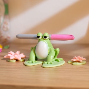 以下が含まれることがあります： 白いお腹と黒い目をした緑色のカエルの形をしたペンホルダーで、グレーとピンクのマーカーを持っています。2つの小さなピンクと緑のスイレンの葉が木製の表面にあります。カエルは退屈そうな表情をしています。