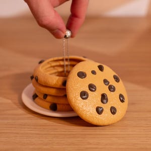 May include: A stack of cookie-shaped tea bag holders with chocolate chip details, on a white plate. A single cookie-shaped tea bag holder is in front of the stack. A hand holds a silver chain attached to the top cookie.
