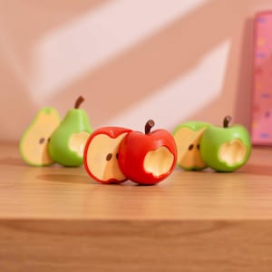 Puede incluir: Colección de figuras de frutas en miniatura, incluyendo manzanas rojas, peras verdes y manzanas verdes, cada una con un mordisco. Las figuras están dispuestas sobre una superficie de madera, con un fondo rosa suave.