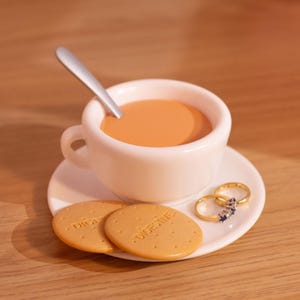 May include: A white teacup and saucer filled with tea, a silver spoon, two biscuits with the word "Digestive" printed on them, and two gold rings with gemstones. The tea is a light brown color.