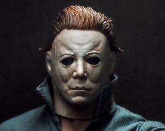 May include: A detailed action figure head and shoulders featuring a pale, expressionless white mask with dark, hollow eye sockets and textured brown hair, wearing a dark blue collared jumpsuit with visible fabric weave and stitching details.