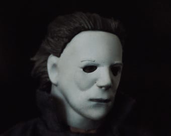 May include: A pale, white, expressionless mask with dark, hollow eye sockets and molded hair, worn over a dark-colored collared garment. The mask features a smooth, matte finish with subtle shading around the eyes and nose, creating a stark contrast against the dark background.