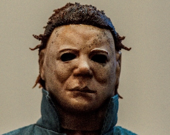 May include: A detailed figurine of a character wearing a weathered, pale, human-like mask with dark, hollow eye sockets and messy brown hair. The figure is dressed in a dark blue collared jumpsuit with a visible texture and a high, stiff collar.