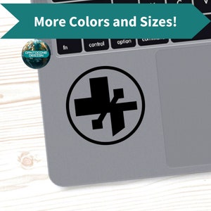 May include: A black sticker featuring a stylized cross and asterisk design within a circular border. The sticker is placed on a gray laptop. The image also includes the text "More Colors and Sizes!" and a logo that says "Dantooine Design".