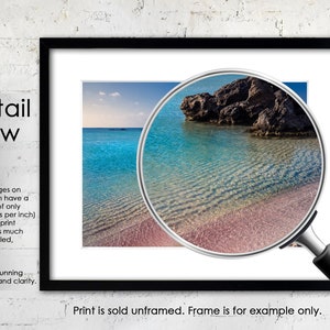 May include: A close-up of a beach with clear turquoise water and a rocky outcrop. The water is shallow and the sand is pink. The image is framed with a black frame and a magnifying glass is over the image.