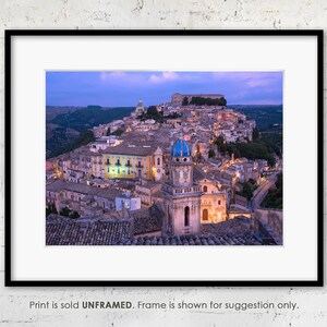 May include: An aerial view of a historic Italian town at dusk. The town is built on a hill and has many buildings with terracotta roofs. A large church with a blue dome is in the center of the image.