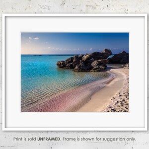 May include: A photograph of a beach with turquoise water and a pink sandy shore. The water is clear and shallow, and there are rocks in the background.