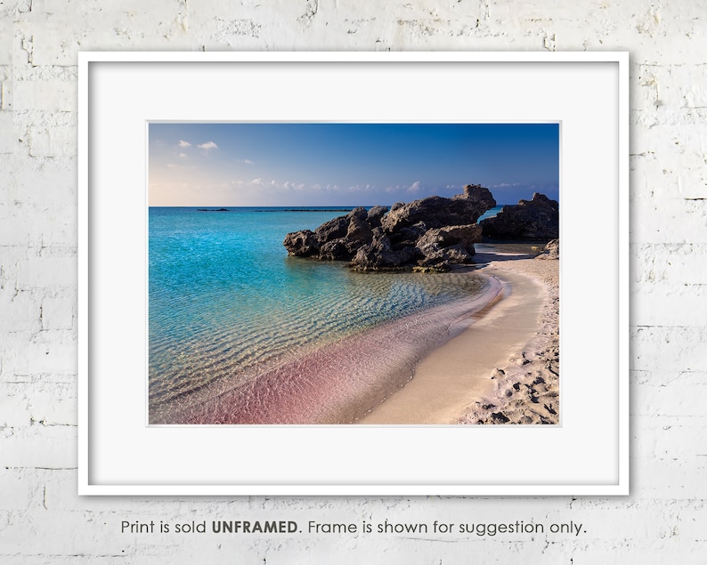May include: A photograph of a beach with turquoise water and a pink sandy shore. The water is clear and shallow, and there are rocks in the background.