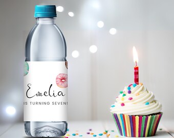 May include: A clear plastic water bottle with a blue lid and a white label that says "Emeliia is turning seven!" with a donut graphic. The bottle is sitting on a white surface with a colorful cupcake with a lit candle.