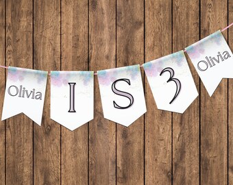 May include: A white banner with a watercolour background and pink, blue, and green accents. The banner says "Olivia is 3" in a purple font.