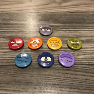 Può includere: Otto spille rotonde con diverse espressioni facciali. Le spille sono in un ordine di colori arcobaleno, a partire dal rosso e terminando con il viola. Le spille sono su una superficie di legno.