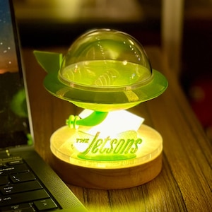 May include: A green and clear plastic flying saucer lamp with a wooden base. The lamp has a clear dome and the words "The Jetsons" are printed on the base.