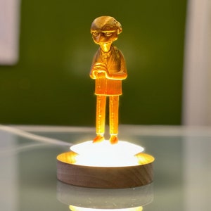 May include: A gold-colored figurine of a man standing on a wooden base with a light shining from underneath. The figurine is wearing a suit and has a serious expression.
